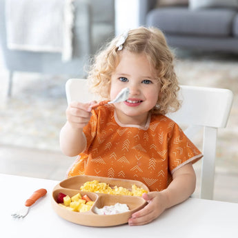 A curly-haired child eagerly eats fruit and scrambled eggs with Bumkins Spoon + Fork: Clay, wearing an orange bib at a white table.
