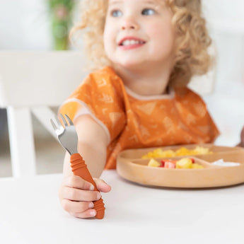 A smiling child in an orange bib holds a Bumkins Spoon + Fork: Clay, sitting at a white table with diced fruit in a brown divided plate.
