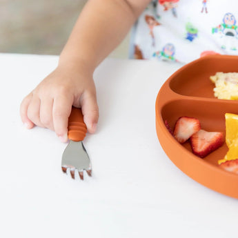 Close-up of a child's hand reaching for a Bumkins Spoon + Fork: Clay on a white table next to a matching plate with strawberries.