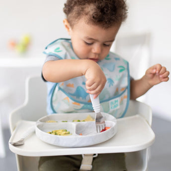 Toddler in high chair uses Bumkins Marble fork to eat from divided plate, wearing dinosaur bib.