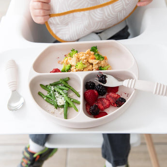 A toddler in a high chair uses Bumkins Spoon + Fork: Sand to eat green beans, berries, and rice with broccoli from a silicone divided plate.