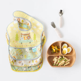 Three colorful bibs with camping designs lay next to Bumkins Spoon + Fork: Sand and a brown divided plate of food on a white background.