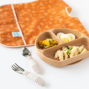 A brown divided plate holds pasta, eggs, and mango on a white background, while Bumkins Spoon + Fork: Sand and an orange bib lay nearby.