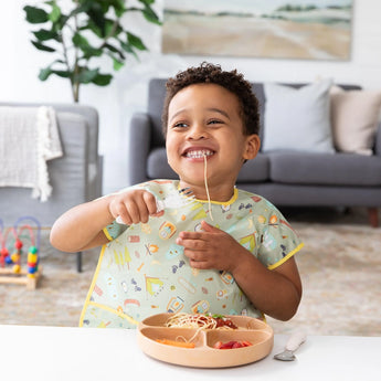A child in a bib, joyfully eating spaghetti with Bumkins Spoon + Fork: Sand from a plate at a white table, in front of a cozy living room.