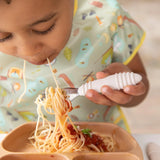 A child eats spaghetti with Bumkins Spoon + Fork: Sand, wearing a patterned bib, using a food-safe brown silicone plate.