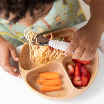 A child eats spaghetti from a brown silicone plate using Bumkins Spoon + Fork: Sand, with strawberries and carrots in the other sections.