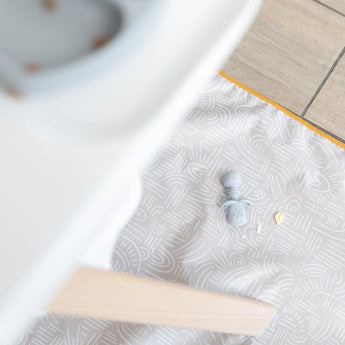 Aerial view of a Bumkins Splat Mat: Wander on a wood floor under a high chair, with a baby spoon and food on top.