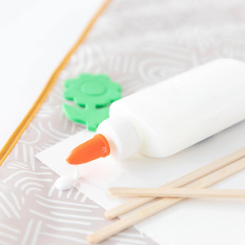Close-up of a leaking white glue bottle with orange tip, green foam flower, wooden sticks, and white paper on a Bumkins Splat Mat: Wander.