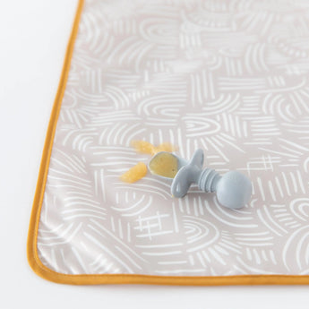 Close-up of a baby spoon with applesauce on a Bumkins Splat Mat: Wander, on a white background.