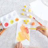 Close-up of a child painting paper on a Bumkins Splat Mat: Wander, surrounded by brushes, a palette, and pom-poms.