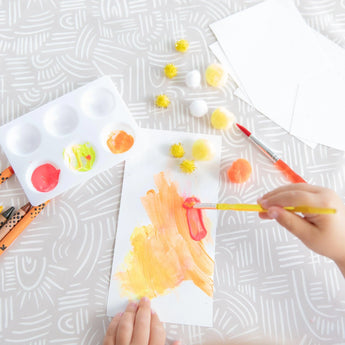 Close-up of a child painting paper on a Bumkins Splat Mat: Wander, surrounded by brushes, a palette, and pom-poms.