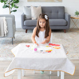 A young girl with a white flower hair clip crafts on a Bumkins Splat Mat: Wander, surrounded by art supplies in a cozy living room.