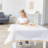 A young girl glues paper on a Bumkins Splat Mat: Wander, surrounded by craft supplies in a bright living room.