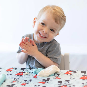 Toddler in grey shirt claps hands on a Bumkins Splat Mat: Mickey Mouse with colorful, waterproof fabric.