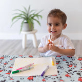 A smiling child with short brown hair sits at a Mickey Mouse-themed table, drawing yellow lines with markers on paper placed on a Bumkins Splat Mat: Mickey Mouse. In the background, a potted plant rests on a white stool.