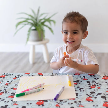 A smiling child with short brown hair sits at a Mickey Mouse-themed table, drawing yellow lines with markers on paper placed on a Bumkins Splat Mat: Mickey Mouse. In the background, a potted plant rests on a white stool.