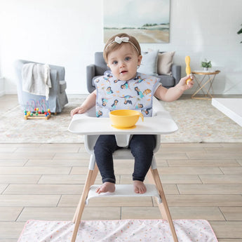 A baby in a high chair with bib, holding a yellow spoon and bowl while Bumkins Splat Mat: Princess Magic covers the floor underneath.