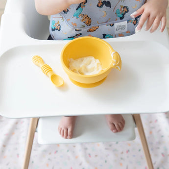 A baby sits in a high chair with a yellow bowl and spoon on the tray, while a blurred Bumkins Splat Mat: Princess Magic lays underneath.