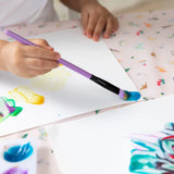 Close-up of a child painting with a purple brush on white paper on a Bumkins Splat Mat: Princess Magic.