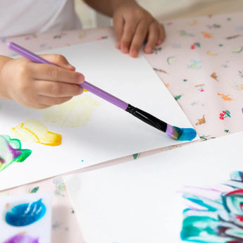 Close-up of a child painting with a purple brush on white paper on a Bumkins Splat Mat: Princess Magic.