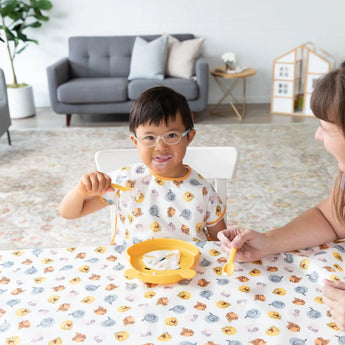 A child with glasses smiles at a table covered with a Bumkins Splat Mat: Winnie and Friends, eating from a plate with an adult nearby.