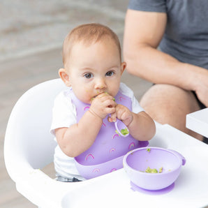 A baby in a high chair wears a Bumkins Silicone Bib: Rainbows, eating with a spoon from a purple bowl.