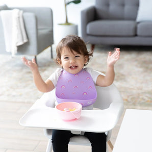 A toddler in a high chair, wearing a Bumkins Silicone Bib: Rainbows, smiles with raised arms with a pink bowl nearby. 