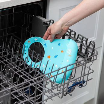 A person places Bumkins Silicone Bib: Ocean Life into the top rack of an open dishwasher.