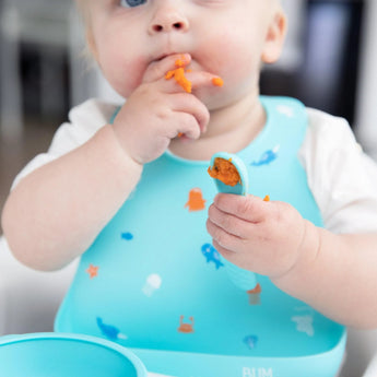 A baby wearing a Bumkins Silicone Bib: Ocean Life eats mashed sweet potato with their hands and a blue spoon.
