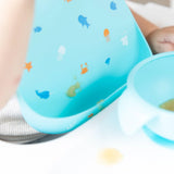 Close-up of a baby in a high chair wearing a Bumkins Silicone Bib: Ocean Life, with focus on a blob of applesauce near the front pocket.