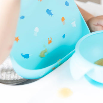 Close-up of a baby in a high chair wearing a Bumkins Silicone Bib: Ocean Life, with focus on a blob of applesauce near the front pocket.