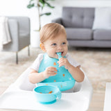 A toddler in a high chair, wearing a Bumkins Silicone Bib: Ocean Life, smiles with spoon in hand with a blue bowl nearby.