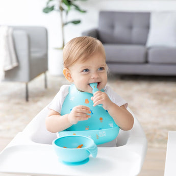 A toddler in a high chair, wearing a Bumkins Silicone Bib: Ocean Life, smiles with spoon in hand with a blue bowl nearby.