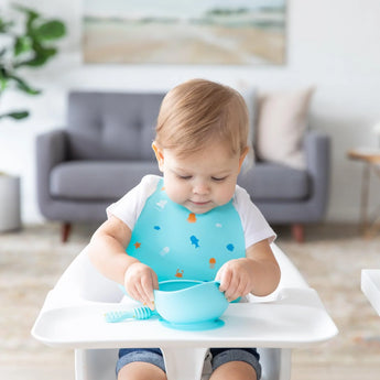 A baby in a high chair enjoys mealtime in a bright room, wearing a Bumkins Silicone Bib: Ocean Life and playing with a blue bowl and spoon.