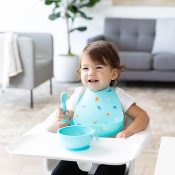 A toddler in a high chair, wearing a Bumkins Silicone Bib: Ocean Life, smiles while holding a spoon with a blue bowl on the tray.