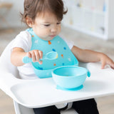 A toddler in a Bumkins Silicone Bib: Ocean Life holds a blue spoon and eyes a blue bowl of food on a high chair tray.