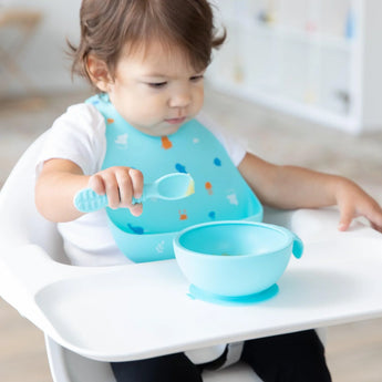 A toddler in a Bumkins Silicone Bib: Ocean Life holds a blue spoon and eyes a blue bowl of food on a high chair tray.