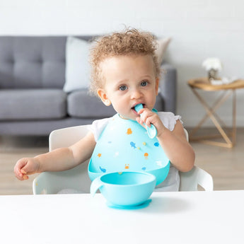 A toddler with curly hair in a high chair wears a Bumkins Silicone Bib: Ocean Life, holding a spoon with a blue bowl in front.