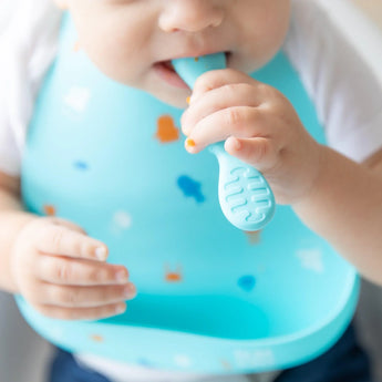 Close-up of a baby wearing a Bumkins Silicone Bib: Ocean Life, chewing on a blue spoon.