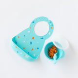 Bumkins Silicone Bib: Ocean Life lays next to a blue bowl with mashed sweet potatoes and a blue spoon on a white background.