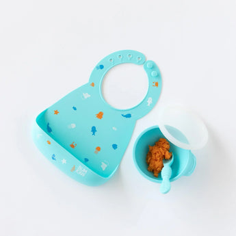 Bumkins Silicone Bib: Ocean Life lays next to a blue bowl with mashed sweet potatoes and a blue spoon on a white background.