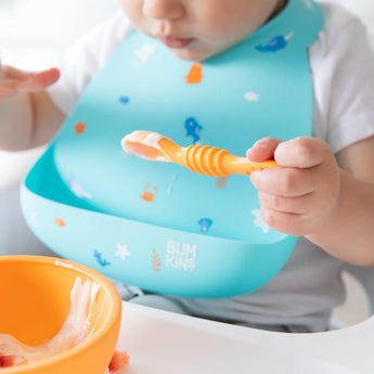 Close-up of a baby sporting a Bumkins Silicone Bib: Ocean Life using an orange spoon and bowl to eat yogurt.