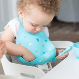 A toddler in a high chair wearing a Bumkins Silicone Bib: Ocean Life reaches into the pocket as they hold a blue spoon.