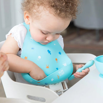 A toddler in a high chair wearing a Bumkins Silicone Bib: Ocean Life reaches into the pocket as they hold a blue spoon.