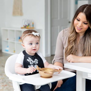 A baby in a high chair in a Bumkins Silicone Bib: Mickey + Minnie Mouse with a gold bowl and spoon is watched by a smiling woman nearby.