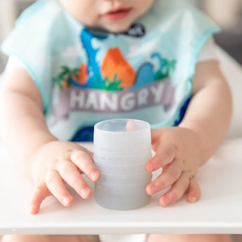 A baby in a high chair wearing a dinosaur bib holds a Bumkins Silicone Starter Cup: Gray.