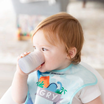 A toddler with light brown hair drinks from a Bumkins Silicone Starter Cup: Gray, wearing a dinosaur-print bib in a softly lit room.