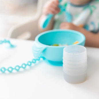 Bumkins Silicone Starter Cup: Gray sits on a white table next to a blue bowl, with a child holding a blue spoon blurred in the background.