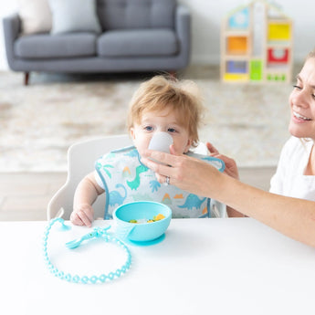 A woman helps a baby in a dinosaur bib drink from a Bumkins Silicone Starter Cup: Gray at a white table with a bowl and spoon.