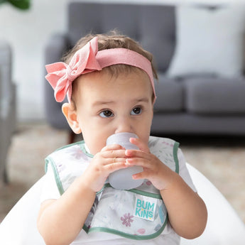 A baby in a pink headband and floral bib drinks from a Bumkins Silicone Starter Cup: Gray in a high chair.